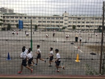 Children are playing and running on a large outdoor playground enclosed by a mesh fence. Various activities are taking place on the asphalt surface, with cones and tires used as part of the setup. Multiple white buildings are visible in the background, indicating a school environment. The area appears spacious and busy, with several groups of students engaging in different games.