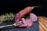 A slicing fork holds up slices of cured sausage on a slate board. Fresh sprigs of rosemary are placed beside the sausage for garnishing. The background is a dark contrast to the rich, reddish hue of the sausages.