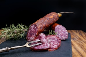 A slicing fork holds up slices of cured sausage on a slate board. Fresh sprigs of rosemary are placed beside the sausage for garnishing. The background is a dark contrast to the rich, reddish hue of the sausages.