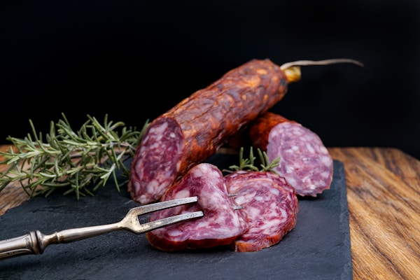 A slicing fork holds up slices of cured sausage on a slate board. Fresh sprigs of rosemary are placed beside the sausage for garnishing. The background is a dark contrast to the rich, reddish hue of the sausages.