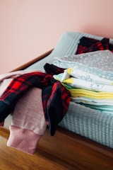 A neatly folded stack of baby clothes placed on a changing table. The clothes include various patterns and colors, with visible prints like cartoon characters. A red and black checkered garment and a pink romper are part of the collection.