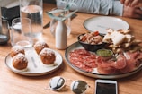 A rustic wooden table is set with an assortment of Italian-style appetizers including round, fried arancini balls topped with cheese on one plate, and a variety of cured meats like prosciutto and salami, alongside pesto and crispy flatbreads on another. Glasses of water, a small white vase with a sprig, and personal items like sunglasses and a smartphone are also visible.
