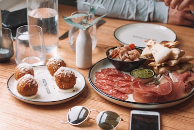 A rustic wooden table is set with an assortment of Italian-style appetizers including round, fried arancini balls topped with cheese on one plate, and a variety of cured meats like prosciutto and salami, alongside pesto and crispy flatbreads on another. Glasses of water, a small white vase with a sprig, and personal items like sunglasses and a smartphone are also visible.