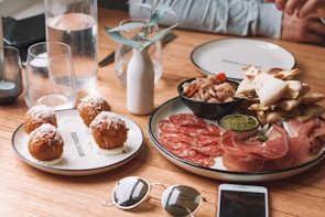 A rustic wooden table is set with an assortment of Italian-style appetizers including round, fried arancini balls topped with cheese on one plate, and a variety of cured meats like prosciutto and salami, alongside pesto and crispy flatbreads on another. Glasses of water, a small white vase with a sprig, and personal items like sunglasses and a smartphone are also visible.