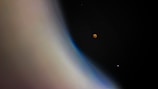 A celestial scene featuring a large planetary body with a smooth gradient of colors ranging from beige to blue, occupying the left side. In the darker expanse of space on the right, two small round objects are visible: one with a reddish hue, resembling a lunar eclipse, and another smaller one with a faint pinkish glow.