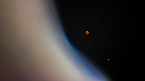 A celestial scene featuring a large planetary body with a smooth gradient of colors ranging from beige to blue, occupying the left side. In the darker expanse of space on the right, two small round objects are visible: one with a reddish hue, resembling a lunar eclipse, and another smaller one with a faint pinkish glow.