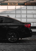 The image depicts the rear side of a sleek black car parked on a cobblestone street. The car's stylish design and glossy finish stand out against the backdrop of a white wall with a sign that reads 'Herbergh Bijsmans' in an ornate font.