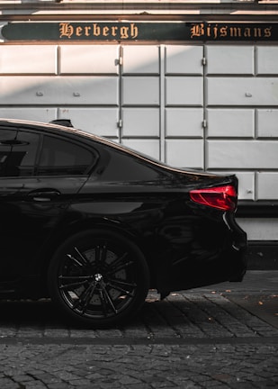 The image depicts the rear side of a sleek black car parked on a cobblestone street. The car's stylish design and glossy finish stand out against the backdrop of a white wall with a sign that reads 'Herbergh Bijsmans' in an ornate font.