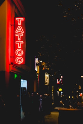 A neon sign reading 'Tattoo' glows brightly in red, hanging on the exterior of a building along a dimly lit street. The street appears to be bustling with a few people walking by, and other illuminated signs and shopfronts are visible in the background, adding to the nighttime urban atmosphere.