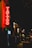 A neon sign reading 'Tattoo' glows brightly in red, hanging on the exterior of a building along a dimly lit street. The street appears to be bustling with a few people walking by, and other illuminated signs and shopfronts are visible in the background, adding to the nighttime urban atmosphere.