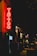 A neon sign reading 'Tattoo' glows brightly in red, hanging on the exterior of a building along a dimly lit street. The street appears to be bustling with a few people walking by, and other illuminated signs and shopfronts are visible in the background, adding to the nighttime urban atmosphere.