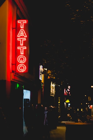 A neon sign reading 'Tattoo' glows brightly in red, hanging on the exterior of a building along a dimly lit street. The street appears to be bustling with a few people walking by, and other illuminated signs and shopfronts are visible in the background, adding to the nighttime urban atmosphere.