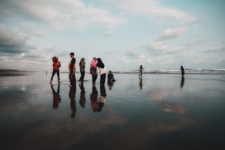 A group of people stands on a wet, reflective beach with the ocean in the background. They are dressed casually, some wearing headscarves, and appear to be interacting with each other. The sky is cloudy, casting a soft light over the scene.