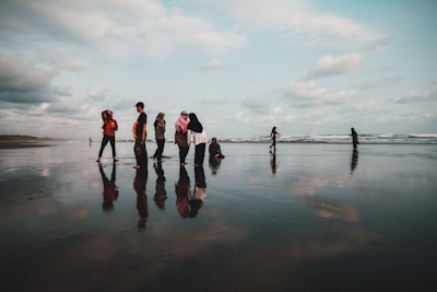 A group of people stands on a wet, reflective beach with the ocean in the background. They are dressed casually, some wearing headscarves, and appear to be interacting with each other. The sky is cloudy, casting a soft light over the scene.