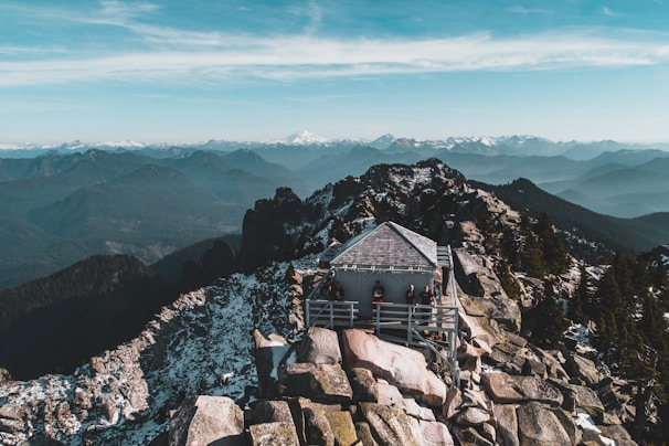 A wooden cabin with a deck is perched on top of a rocky mountain peak. The surrounding landscape features a vast expanse of mountain ranges stretching into the distance, partially covered in snow. The sky is clear with a few wispy clouds, and there are several people gathered on the deck, enjoying the view.