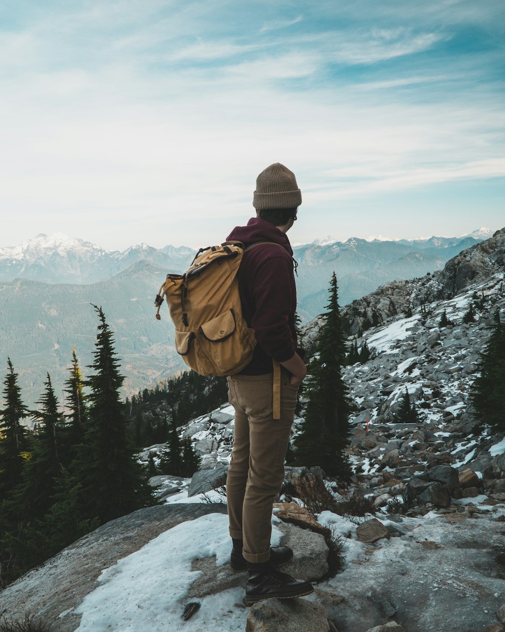 person wearing jacket and hiking backpack standing on cliff while watching mountain during daytime