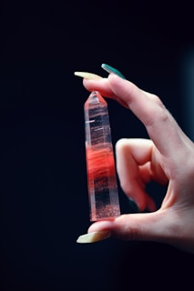 A hand with long nails delicately holds a polished, red gemstone that is pointed at one end. The gemstone has visible layers and appears translucent, with reflections catching the light. The background is dark, emphasizing the colors and details of the hand and stone.