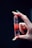 A hand with long nails delicately holds a polished, red gemstone that is pointed at one end. The gemstone has visible layers and appears translucent, with reflections catching the light. The background is dark, emphasizing the colors and details of the hand and stone.