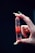 A hand with long nails delicately holds a polished, red gemstone that is pointed at one end. The gemstone has visible layers and appears translucent, with reflections catching the light. The background is dark, emphasizing the colors and details of the hand and stone.