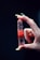 A hand with long nails delicately holds a polished, red gemstone that is pointed at one end. The gemstone has visible layers and appears translucent, with reflections catching the light. The background is dark, emphasizing the colors and details of the hand and stone.