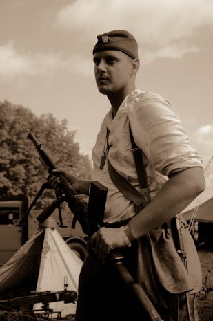 A person wearing a vintage military uniform and hat, holding a rifle, stands in a campsite with a tent and military vehicle visible in the background. The scene is in sepia tones, suggesting a historical or reenactment setting.