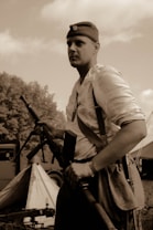 A person wearing a vintage military uniform and hat, holding a rifle, stands in a campsite with a tent and military vehicle visible in the background. The scene is in sepia tones, suggesting a historical or reenactment setting.