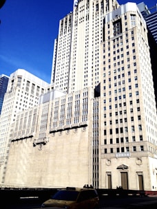 Tall, beige skyscraper against a clear blue sky with decorative architectural details and large windows. A yellow taxi is visible in the foreground, adding a touch of everyday life to the urban scene.
