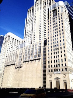 Tall, beige skyscraper against a clear blue sky with decorative architectural details and large windows. A yellow taxi is visible in the foreground, adding a touch of everyday life to the urban scene.