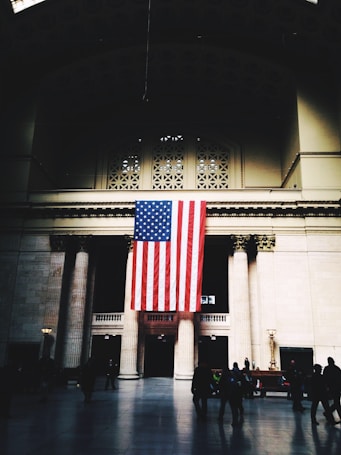 A large American flag hangs prominently in the center of a grand hall with high ceilings and tall columns. The space is dimly lit, with patches of light creating a dramatic atmosphere. People are scattered in the foreground, adding a sense of activity to the scene.