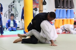 Two individuals are practicing martial arts in a gym setting. They are wearing traditional martial arts uniforms, known as gis, and are engaged in grappling on a padded floor. In the background, others in similar attire are seated or standing, watching or preparing for their turn. Various training equipment and padded shields are visible, suggesting this is a martial arts or jiu-jitsu class.
