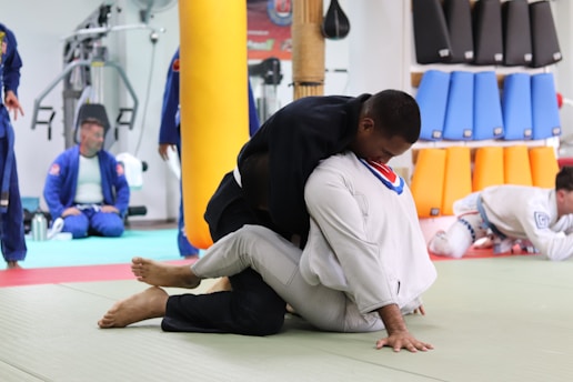 Two individuals are practicing martial arts in a gym setting. They are wearing traditional martial arts uniforms, known as gis, and are engaged in grappling on a padded floor. In the background, others in similar attire are seated or standing, watching or preparing for their turn. Various training equipment and padded shields are visible, suggesting this is a martial arts or jiu-jitsu class.