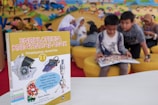 A children's encyclopedia about creativity is prominently displayed. The book features illustrations of space-related technology such as a satellite, space capsule, spacesuit, rocket, and a spectrograph on its cover. In the background, children are sitting on yellow cushioned seats, reading and engaging with each other. The wall behind them is decorated with a colorful mural of a cartoon-like scene.