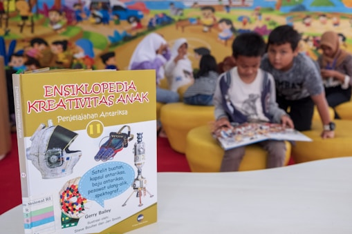 A children's encyclopedia about creativity is prominently displayed. The book features illustrations of space-related technology such as a satellite, space capsule, spacesuit, rocket, and a spectrograph on its cover. In the background, children are sitting on yellow cushioned seats, reading and engaging with each other. The wall behind them is decorated with a colorful mural of a cartoon-like scene.
