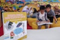 A children's encyclopedia about creativity is prominently displayed. The book features illustrations of space-related technology such as a satellite, space capsule, spacesuit, rocket, and a spectrograph on its cover. In the background, children are sitting on yellow cushioned seats, reading and engaging with each other. The wall behind them is decorated with a colorful mural of a cartoon-like scene.