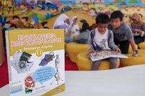 A children's encyclopedia about creativity is prominently displayed. The book features illustrations of space-related technology such as a satellite, space capsule, spacesuit, rocket, and a spectrograph on its cover. In the background, children are sitting on yellow cushioned seats, reading and engaging with each other. The wall behind them is decorated with a colorful mural of a cartoon-like scene.