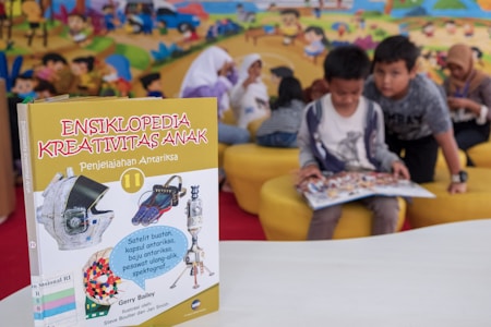 A children's encyclopedia about creativity is prominently displayed. The book features illustrations of space-related technology such as a satellite, space capsule, spacesuit, rocket, and a spectrograph on its cover. In the background, children are sitting on yellow cushioned seats, reading and engaging with each other. The wall behind them is decorated with a colorful mural of a cartoon-like scene.