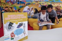 A children's encyclopedia about creativity is prominently displayed. The book features illustrations of space-related technology such as a satellite, space capsule, spacesuit, rocket, and a spectrograph on its cover. In the background, children are sitting on yellow cushioned seats, reading and engaging with each other. The wall behind them is decorated with a colorful mural of a cartoon-like scene.