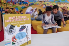 A children's encyclopedia about creativity is prominently displayed. The book features illustrations of space-related technology such as a satellite, space capsule, spacesuit, rocket, and a spectrograph on its cover. In the background, children are sitting on yellow cushioned seats, reading and engaging with each other. The wall behind them is decorated with a colorful mural of a cartoon-like scene.