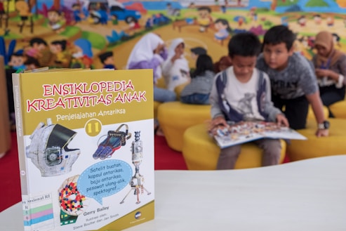 A children's encyclopedia about creativity is prominently displayed. The book features illustrations of space-related technology such as a satellite, space capsule, spacesuit, rocket, and a spectrograph on its cover. In the background, children are sitting on yellow cushioned seats, reading and engaging with each other. The wall behind them is decorated with a colorful mural of a cartoon-like scene.