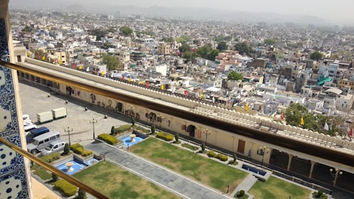 A sprawling urban landscape with a densely packed array of buildings and flat rooftops. In the foreground, there is a well-maintained area featuring manicured lawns, small fountains, and pathways. A decorative wall with flags lines the area, separating the gardens from the cityscape. The hazy sky blends with the distant hills on the horizon.