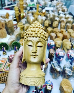 A hand holds a detailed wooden sculpture of a serene face with closed eyes and intricate hair patterns. The background is filled with various small wooden crafts and decorative items displayed on a white table.