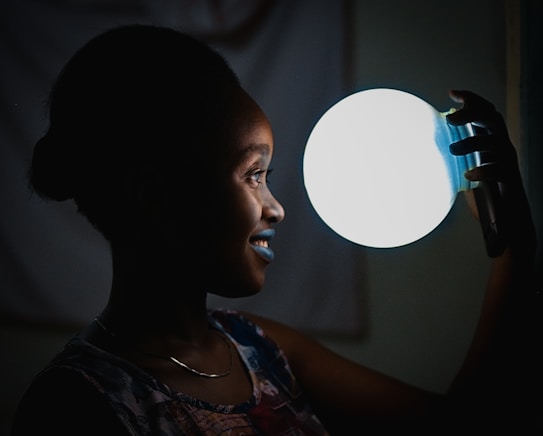 A person with a joyful expression holds a bright, circular light source close to their face in a dimly lit setting. The contrast between the darkness and the glow creates a visually striking effect, highlighting the details on the person's profile.