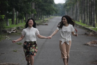 Two people are joyfully holding hands while walking down a tree-lined pathway. They appear to be enjoying themselves, each holding a small bunch of flowers. The atmosphere is light and carefree, enhanced by the greenery on either side of the path.