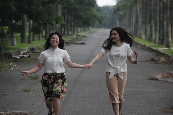 Two people are joyfully holding hands while walking down a tree-lined pathway. They appear to be enjoying themselves, each holding a small bunch of flowers. The atmosphere is light and carefree, enhanced by the greenery on either side of the path.