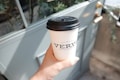 A hand holding a white disposable coffee cup with a black plastic lid, featuring the branding of a coffee roastery. The background includes a light blue door with brass handles and some greenery, suggesting an outdoor setting or near a cafe entrance.