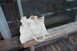 A beige cloth tote bag with the word 'VOLUNTEER' printed on it, placed on a rustic wooden bench. The background features a glass wall with a grid pattern, possibly a window or a door, offering a view of a brick wall.