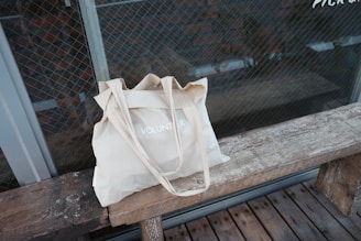 A beige cloth tote bag with the word 'VOLUNTEER' printed on it, placed on a rustic wooden bench. The background features a glass wall with a grid pattern, possibly a window or a door, offering a view of a brick wall.