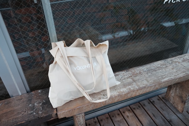 A beige cloth tote bag with the word 'VOLUNTEER' printed on it, placed on a rustic wooden bench. The background features a glass wall with a grid pattern, possibly a window or a door, offering a view of a brick wall.