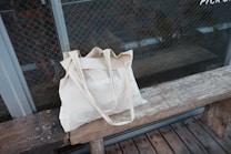 A beige cloth tote bag with the word 'VOLUNTEER' printed on it, placed on a rustic wooden bench. The background features a glass wall with a grid pattern, possibly a window or a door, offering a view of a brick wall.