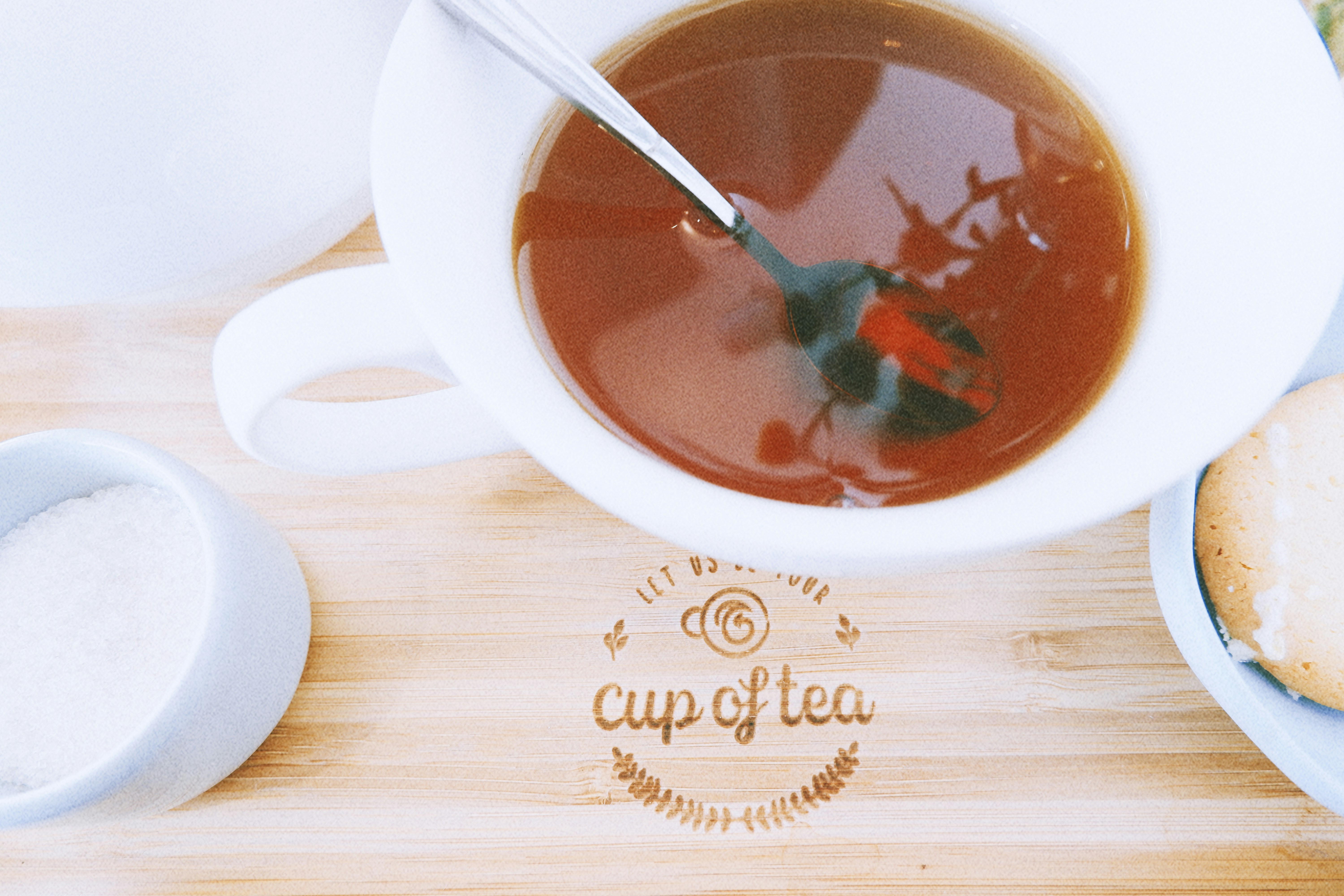 A photograph of a white teacup with tea on a light wood tray, centered around a carved 'cup of tea' logo. Left is a small sugar bowl and right a biscuit plate.
